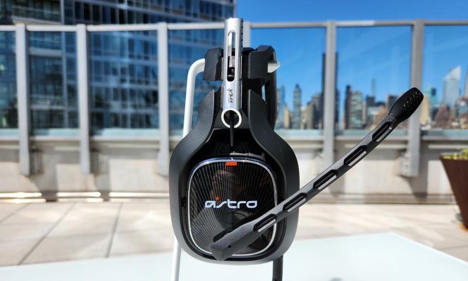 O fone de ouvido Astro A40 TR Gaming repousa em um suporte branco em uma mesa externa, com seu microfone embutido estendido.