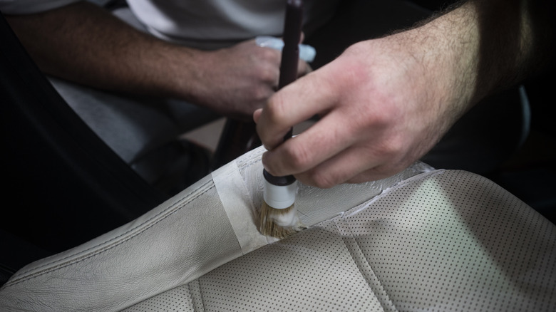 Mão segurando uma escova de detalhamento limpando um assento de carro de couro bege perfurado