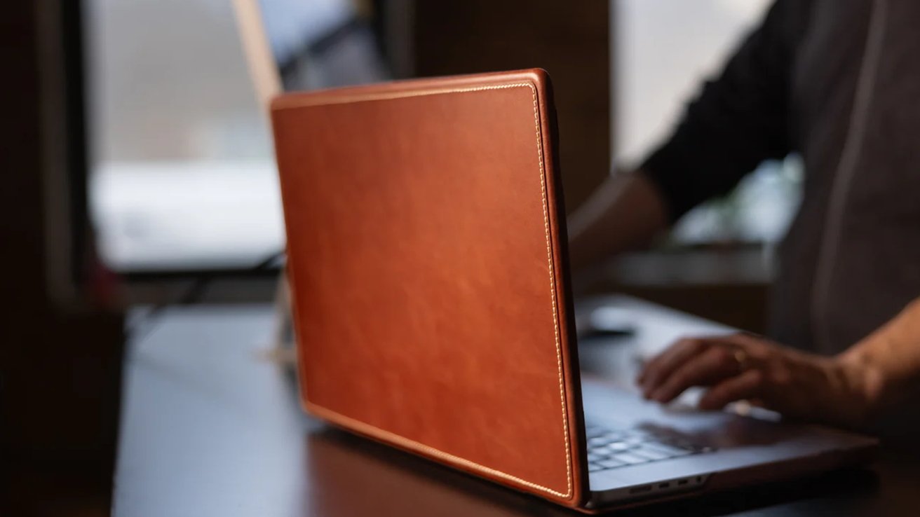 Pessoa digitando em um laptop com capa de couro marrom em uma sala com iluminação suave.