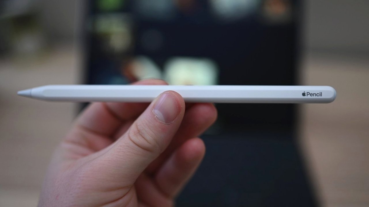 Close de uma mão segurando um Apple Pencil em foco, com um fundo desfocado mostrando um tablet.