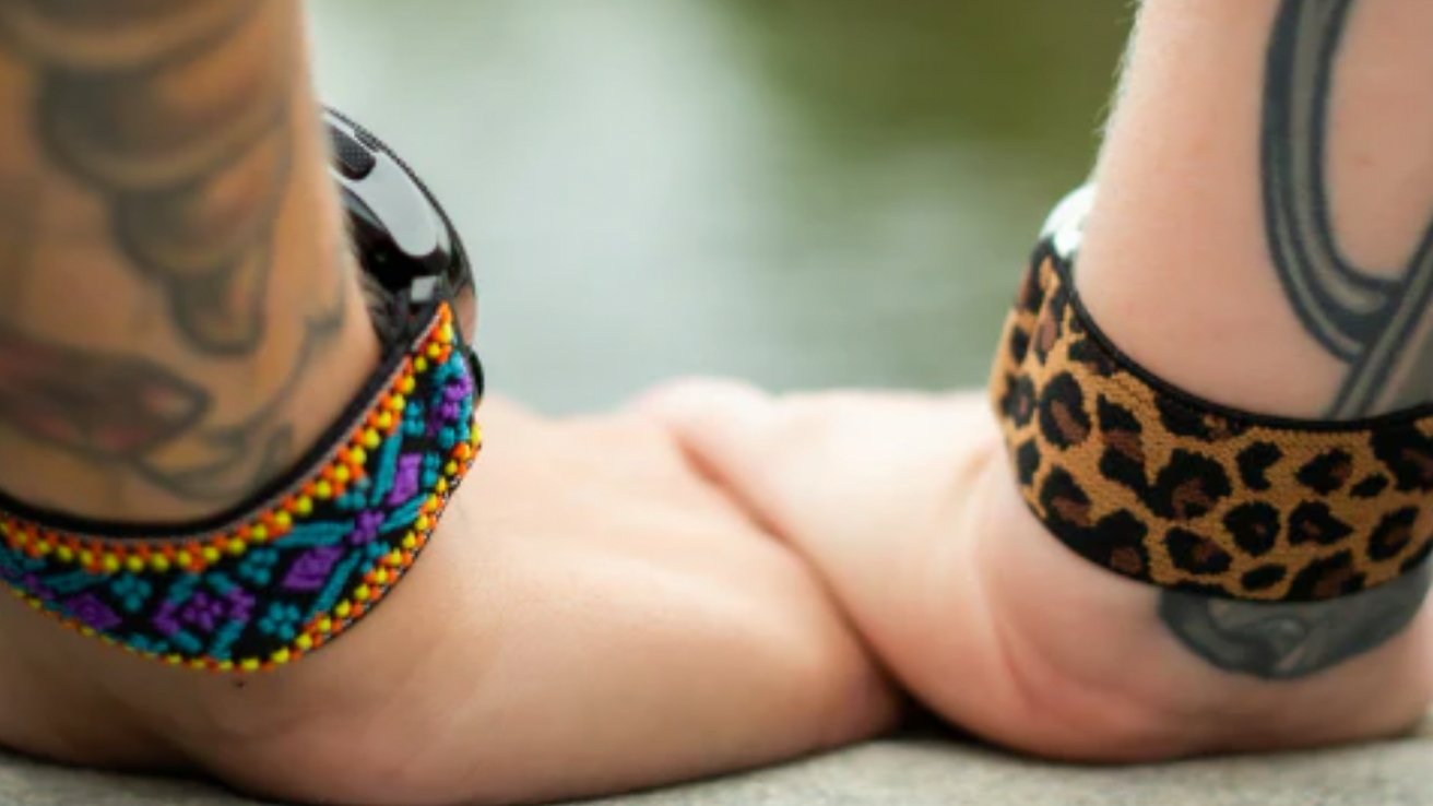 Duas mãos com tatuagens, usando pulseiras de relógio coloridas, apoiadas juntas contra um fundo desfocado ao ar livre.