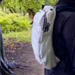 The Outdoor Backpack worn outside in profile next to a damp tree with lots of green foliage in the background.