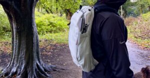 The Outdoor Backpack worn outside in profile next to a damp tree with lots of green foliage in the background.