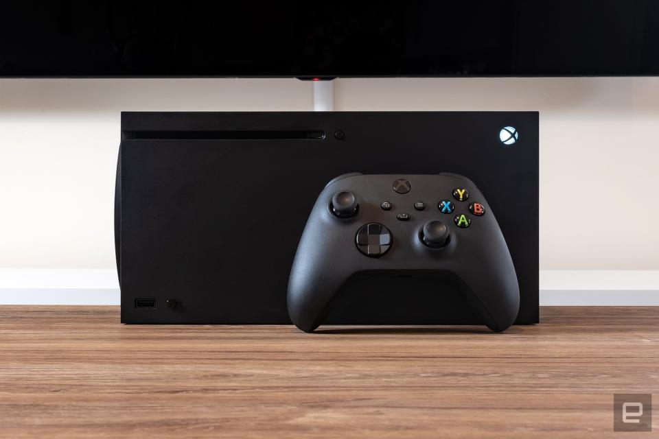 O console de jogos Xbox Series X está deitado de lado, com um controle Xbox preto apoiado na frente dele, em cima de uma mesa de madeira marrom.