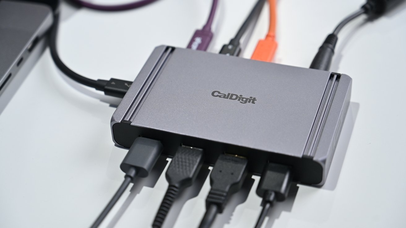 Um hub CalDigit compacto com vários cabos conectados, incluindo USB e HDMI, ao lado de um laptop em uma superfície branca.