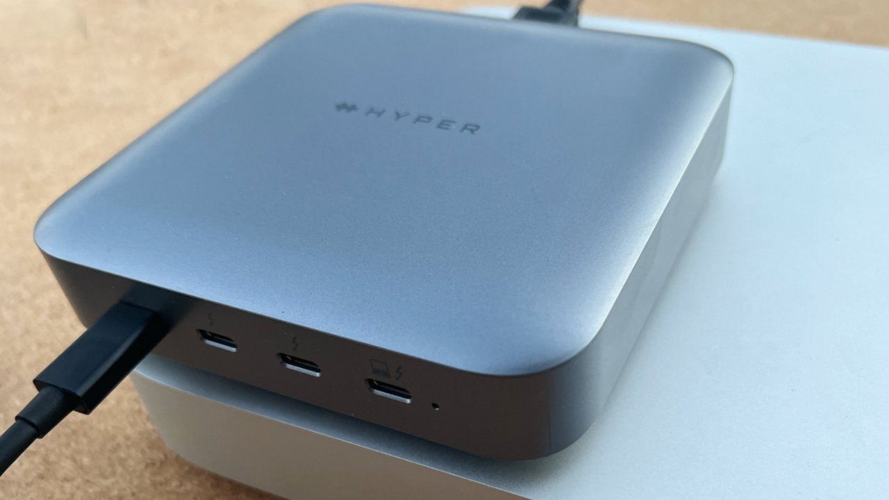 Dispositivo eletrônico prateado compacto com a marca 'Hyper', com três portas USB-C e um cabo conectado, colocado em uma superfície branca.