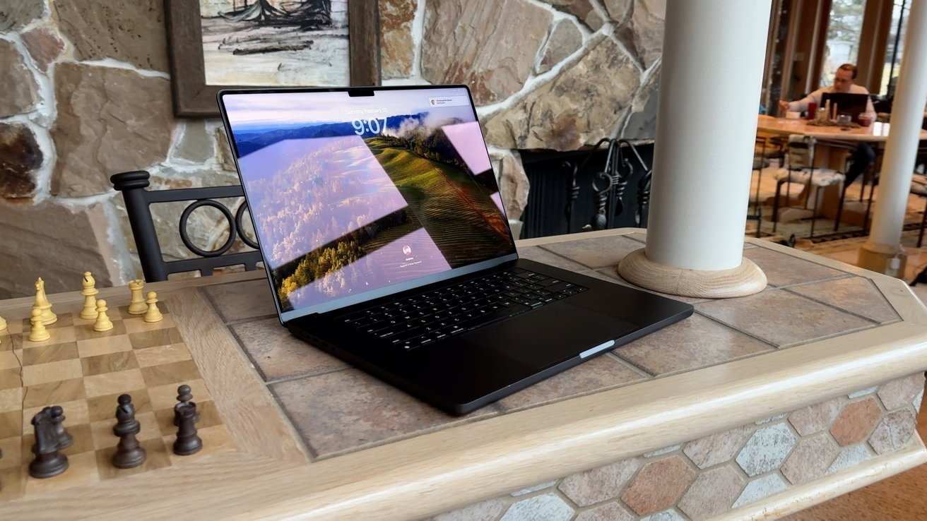 Um MacBook Pro de 16 polegadas exibindo uma foto cênica de uma paisagem fica em um balcão de pedra perto de um tabuleiro de xadrez com uma lareira de pedra ao fundo.