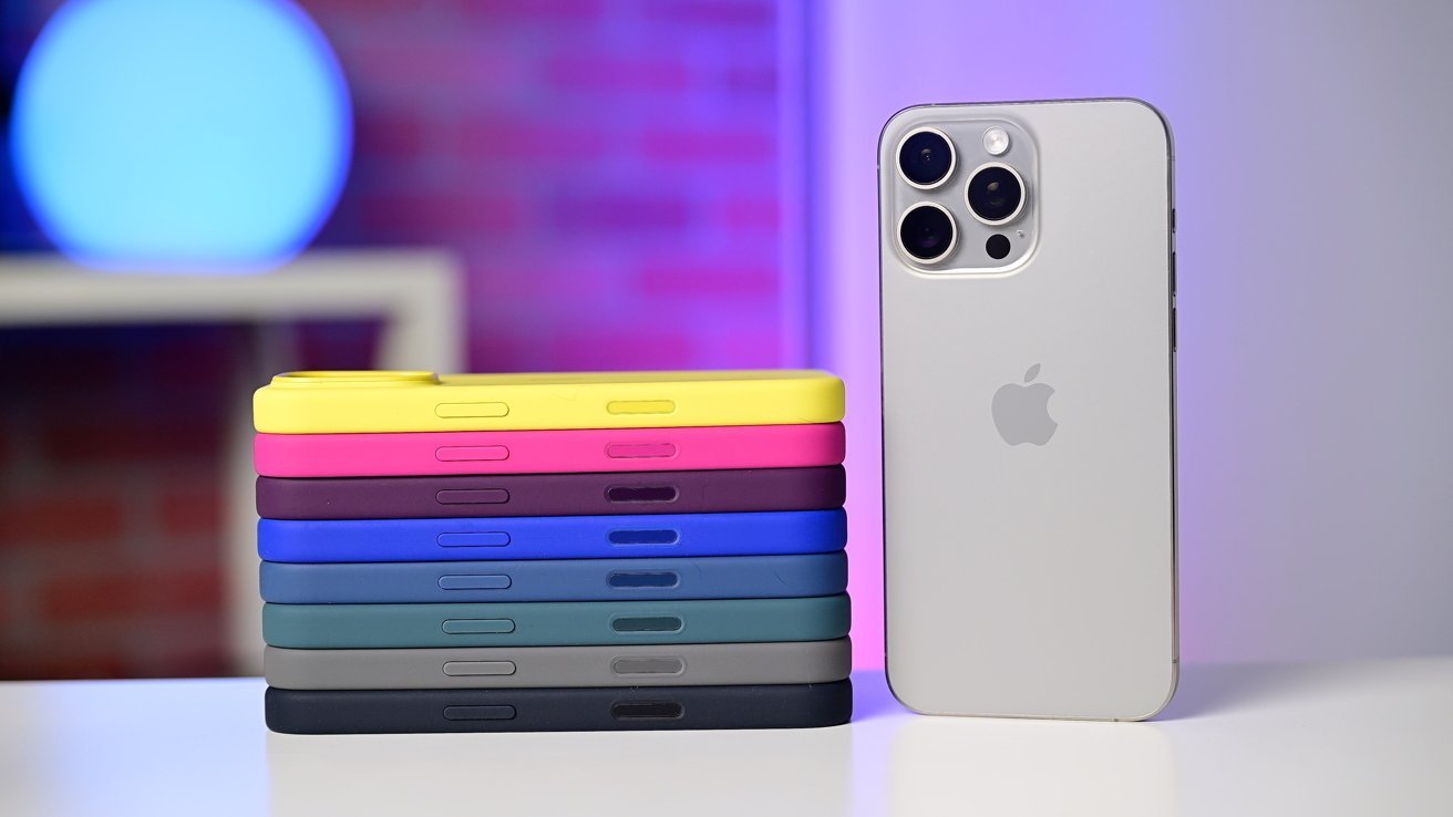 O Apple iPhone 16 Pro fica ao lado de uma pilha de capas coloridas de silicone da Apple, incluindo amarelo, rosa, vermelho e tons de azul e verde. O fundo tem um brilho azul e roxo.