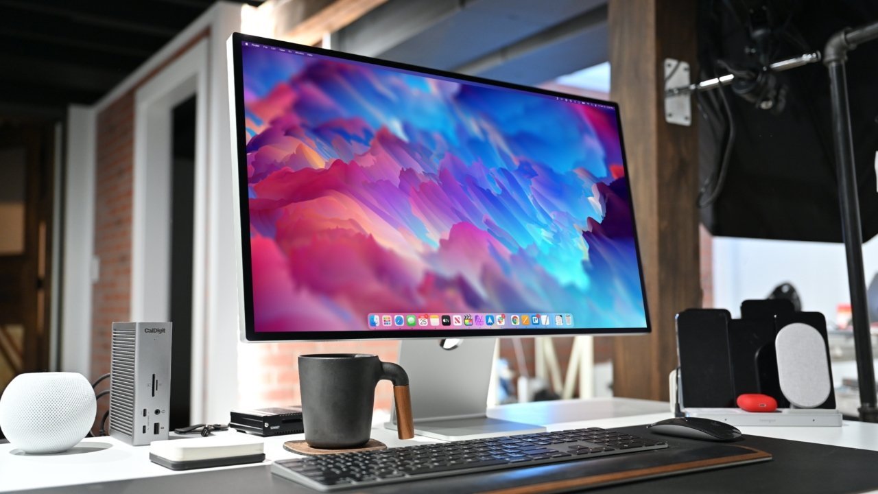 Um computador elegante com um grande Apple Studio Display, teclado preto, caneca de café e acessórios em uma mesa branca moderna.