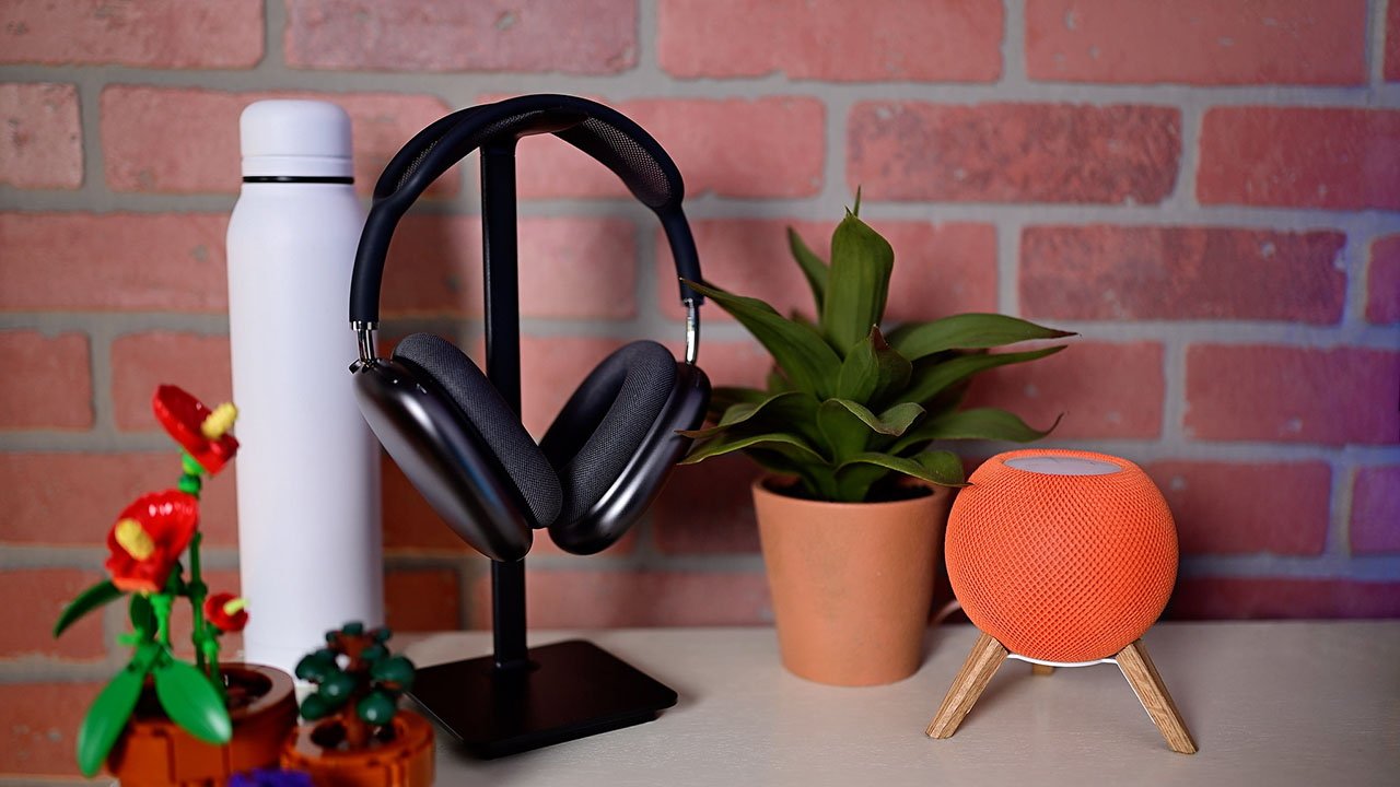 AirPods Max no suporte, garrafa branca, vaso de planta, mini alto-falante HomePod laranja e flores de brinquedo na mesa contra o fundo da parede de tijolos.