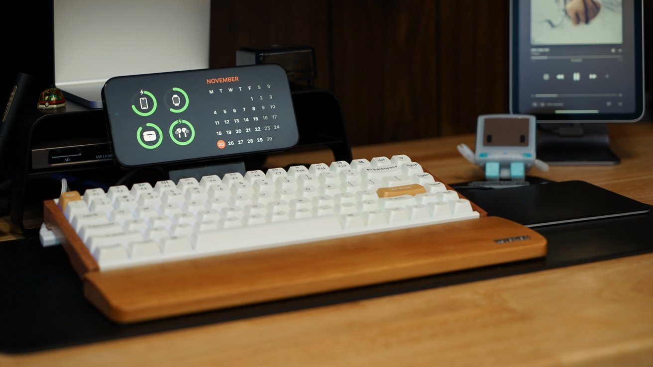 Configuração de mesa com teclado branco, telefone exibindo calendário, pequena figura de robô e tablet tocando música, tudo em uma superfície de madeira.