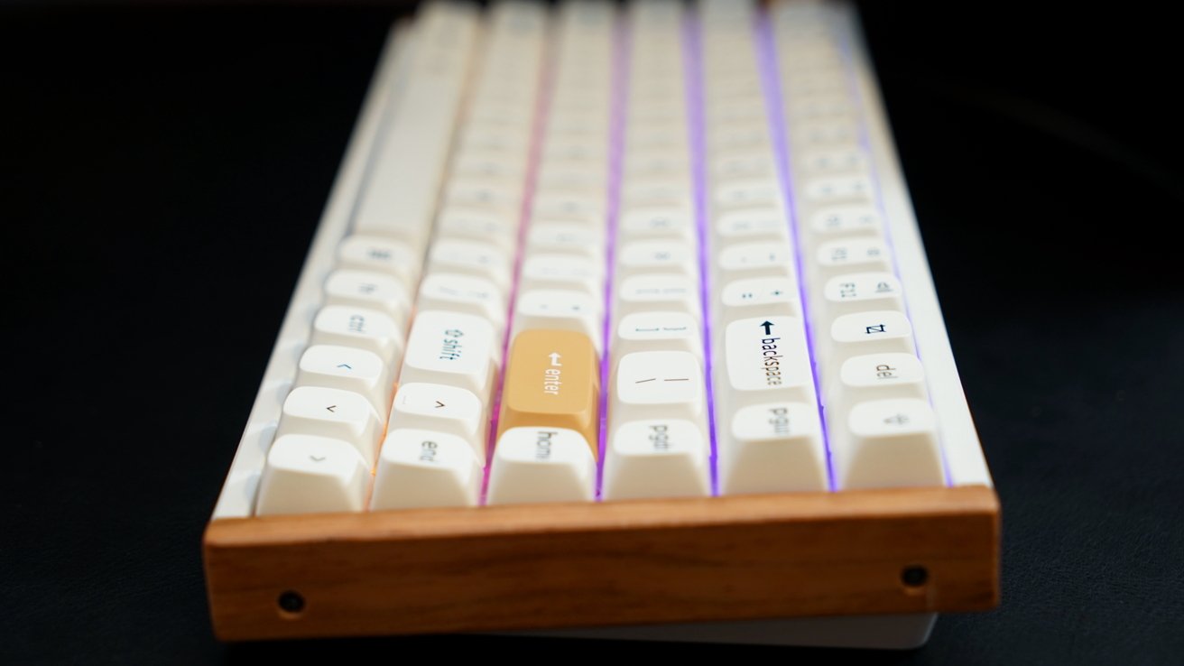 Teclado mecânico branco com retroiluminação roxa, com pousa palmas de madeira e tecla laranja entre as teclas brancas.