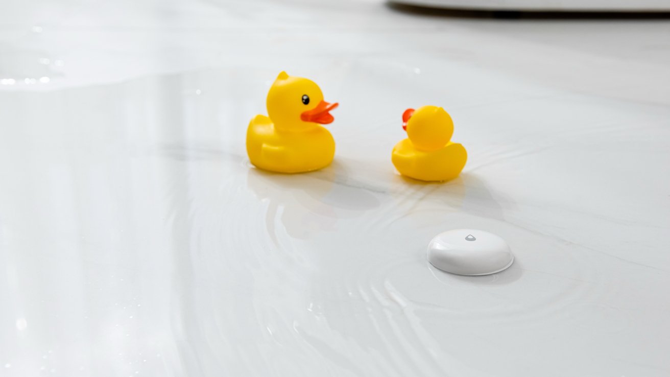 Dois patos de borracha amarelos sobre uma superfície branca molhada, ao lado de um pequeno dispositivo circular branco com um símbolo de gota de água.