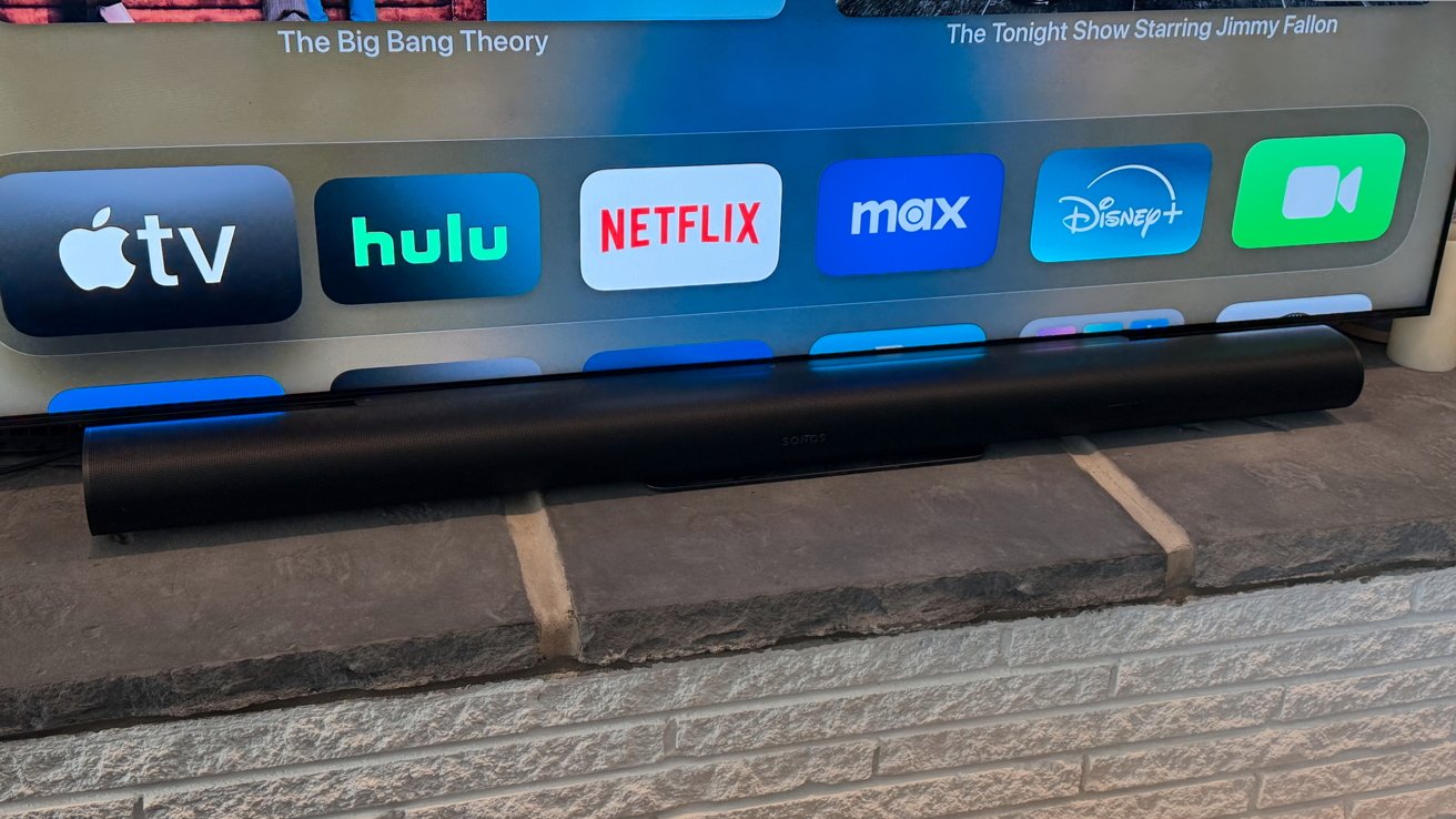 Tela de televisão exibindo aplicativos de streaming como Apple TV, Hulu, Netflix e Disney+ acima de uma barra de som Sonos preta em uma superfície de pedra.