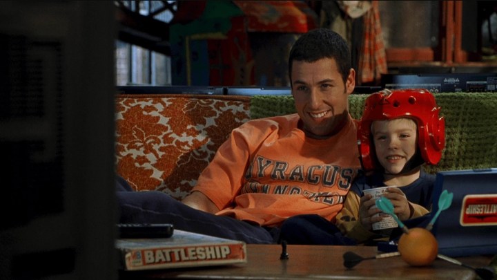 Adam Sandler abraçando um menino de capacete, ambos assistindo TV em uma cena de Big Daddy.