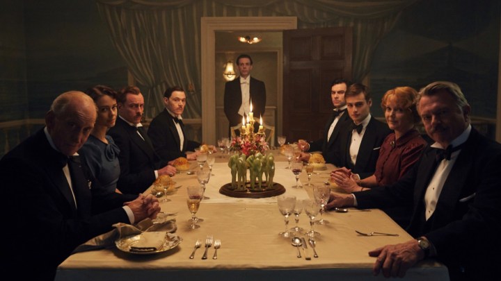 Convidados sentados em uma mesa de jantar olham para a câmera em And Then There Were None.