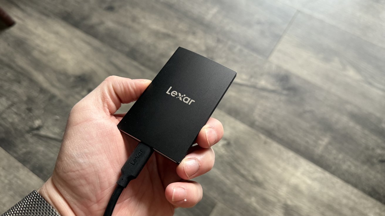 Mão segurando um SSD externo Lexar preto em um piso cinza com textura de madeira.