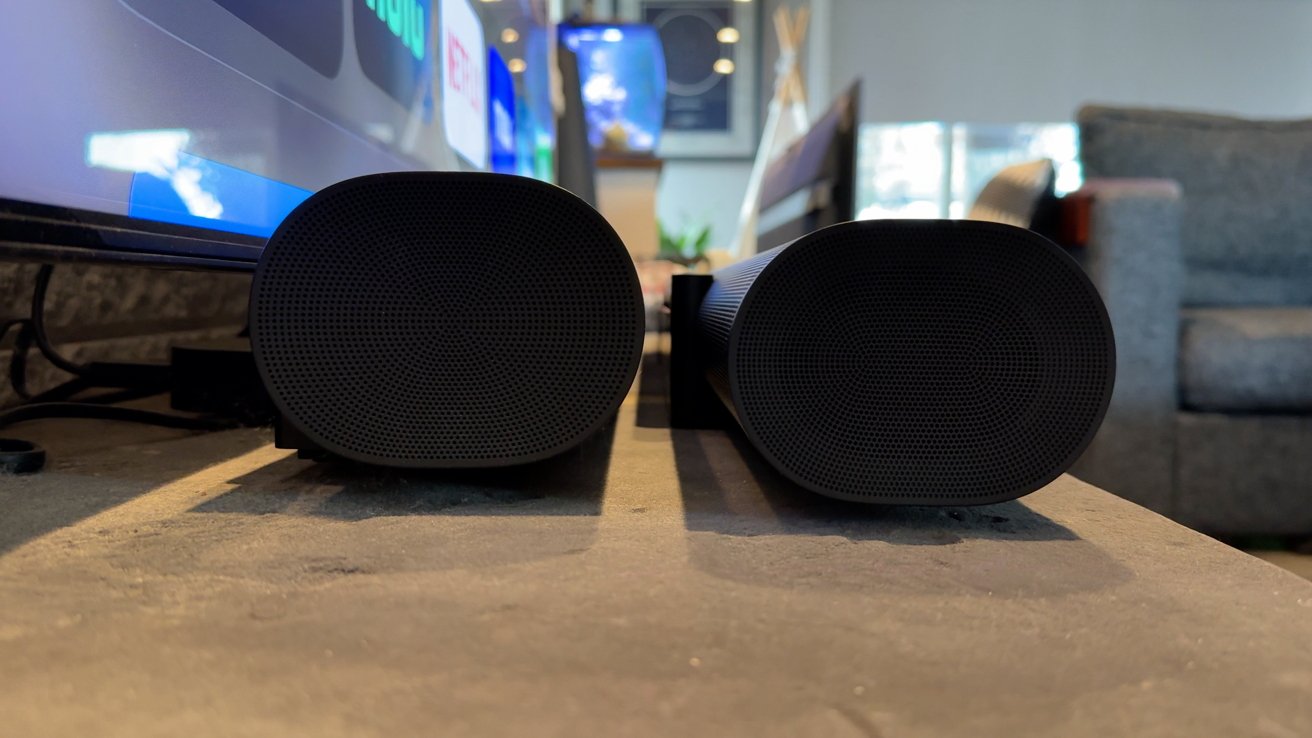 Dois alto-falantes cilíndricos pretos lado a lado sobre uma mesa em uma sala de estar, com uma televisão e um sofá ao fundo.