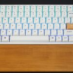 Teclado mecânico branco com pousa palmas de madeira, com teclas escape e enter laranja, colocado sobre superfície escura.