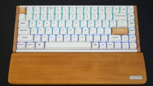 Teclado mecânico branco com pousa palmas de madeira, com teclas escape e enter laranja, colocado sobre superfície escura.