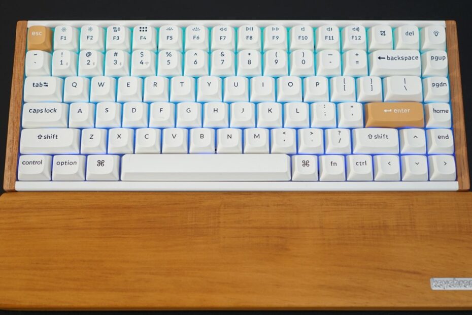 Teclado mecânico branco com pousa palmas de madeira, com teclas escape e enter laranja, colocado sobre superfície escura.