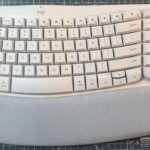 Teclado ergonômico branco com teclas curvas e pousa palmas cinza, colocado em uma superfície com padrão de grade.