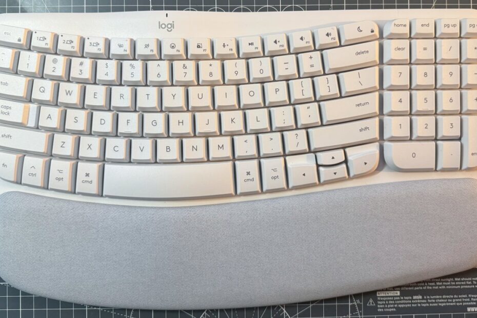 Teclado ergonômico branco com teclas curvas e pousa palmas cinza, colocado em uma superfície com padrão de grade.