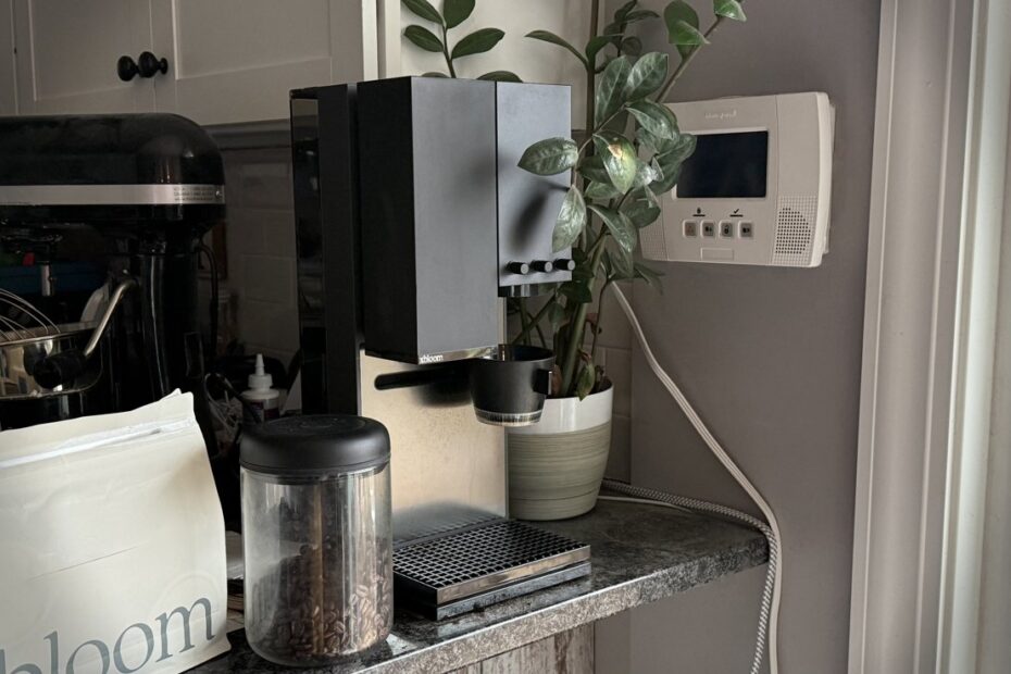 Máquina de café na bancada da cozinha com um vaso de planta, grãos de café em uma jarra, batedeira e termostato de parede próximos.