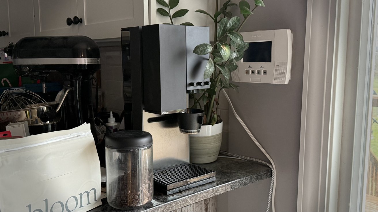 Máquina de café na bancada da cozinha com um vaso de planta, grãos de café em uma jarra, batedeira e termostato de parede próximos.