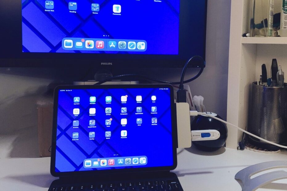 Um iPad conectado a um teclado em uma mesa, exibindo uma tela azul com ícones de aplicativos, espelhados em um monitor montado na parede. Prateleiras adjacentes armazenam materiais de escritório e produtos de limpeza.