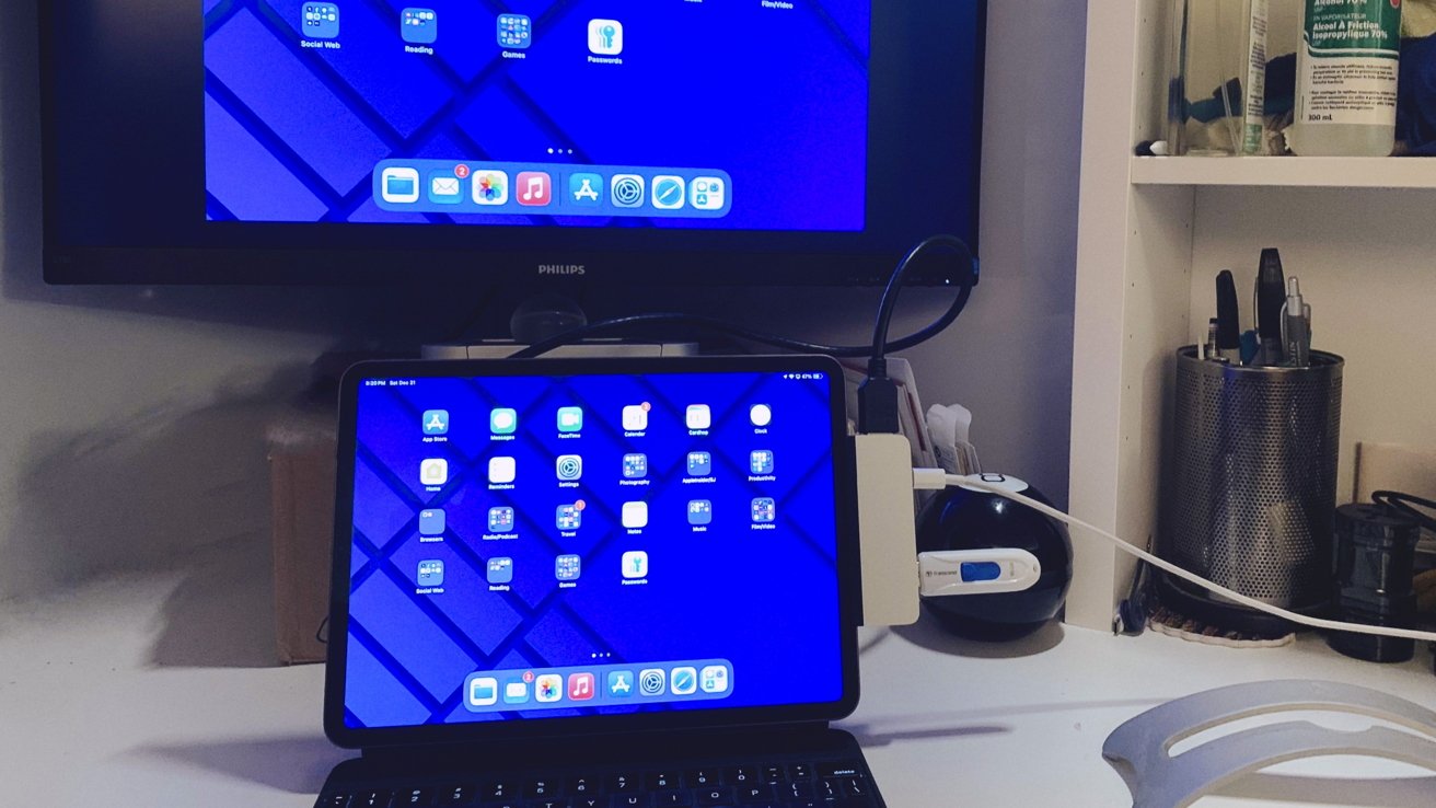 Um iPad conectado a um teclado em uma mesa, exibindo uma tela azul com ícones de aplicativos, espelhados em um monitor montado na parede. Prateleiras adjacentes armazenam materiais de escritório e produtos de limpeza.