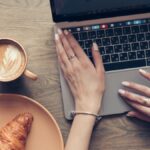 Mãos no teclado do MacBook ao lado de um café e um croissant.