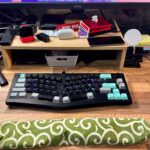 Uma mesa organizada com teclado, mouse, vários gadgets, um smartphone em um suporte e um pousa palmas com padrão verde em uma superfície de madeira.