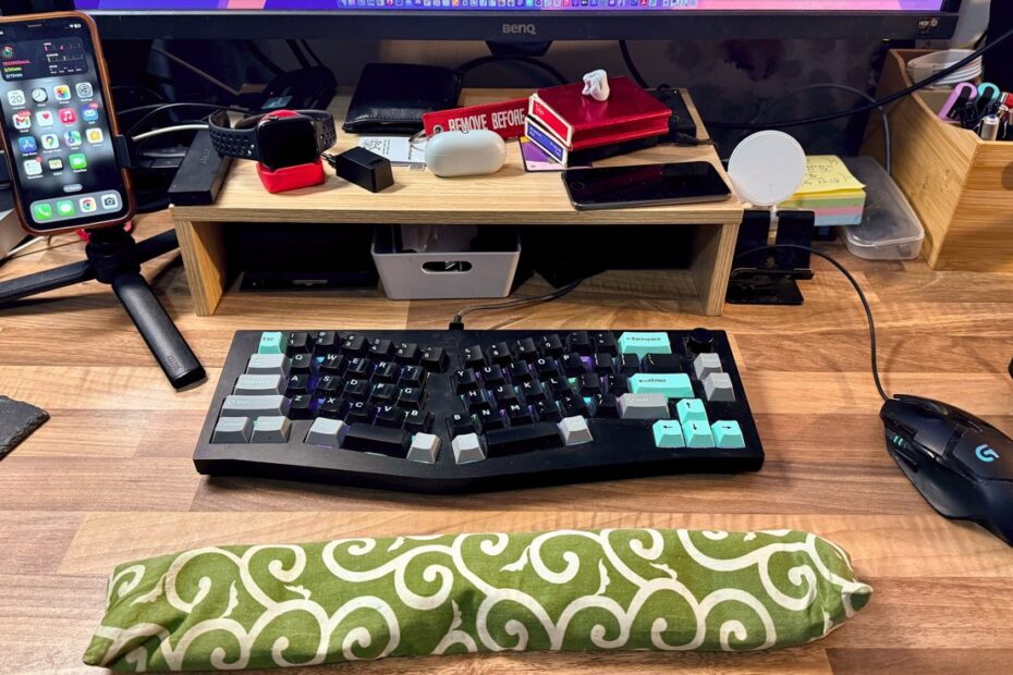 Uma mesa organizada com teclado, mouse, vários gadgets, um smartphone em um suporte e um pousa palmas com padrão verde em uma superfície de madeira.