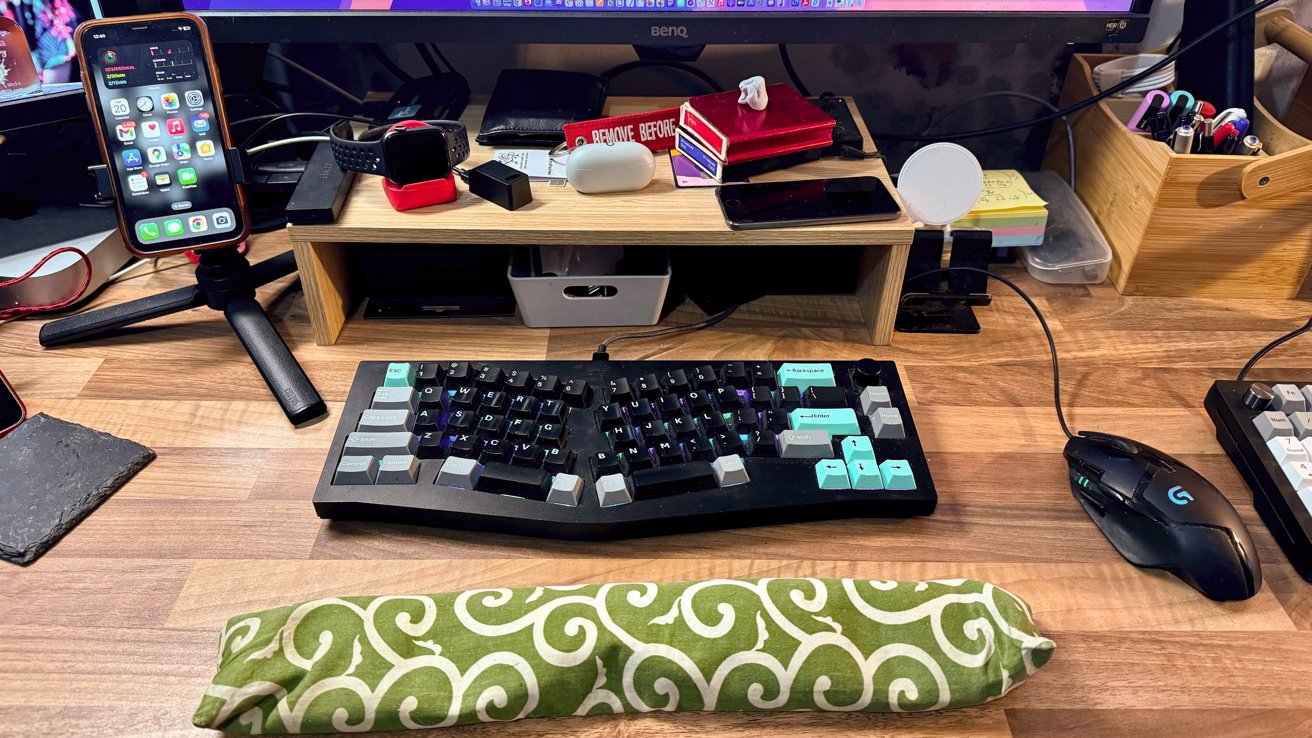 Uma mesa organizada com teclado, mouse, vários gadgets, um smartphone em um suporte e um pousa palmas com padrão verde em uma superfície de madeira.