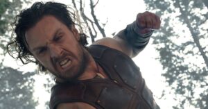 A muscular man in a leather vest leaning down to punch someone in a forest.