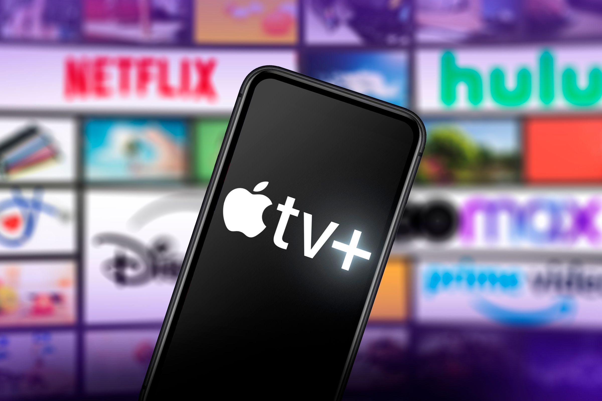 Um telefone com o logotipo da Apple TV+ na tela e ao fundo diversas telas com logotipos de outros serviços de streaming.