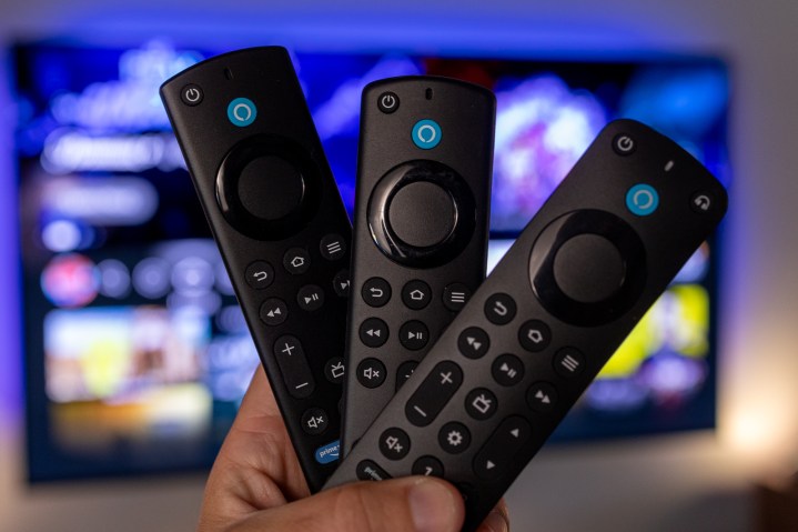 Um trio de controles remotos da Amazon Fire TV segurados na mão em frente a uma televisão.