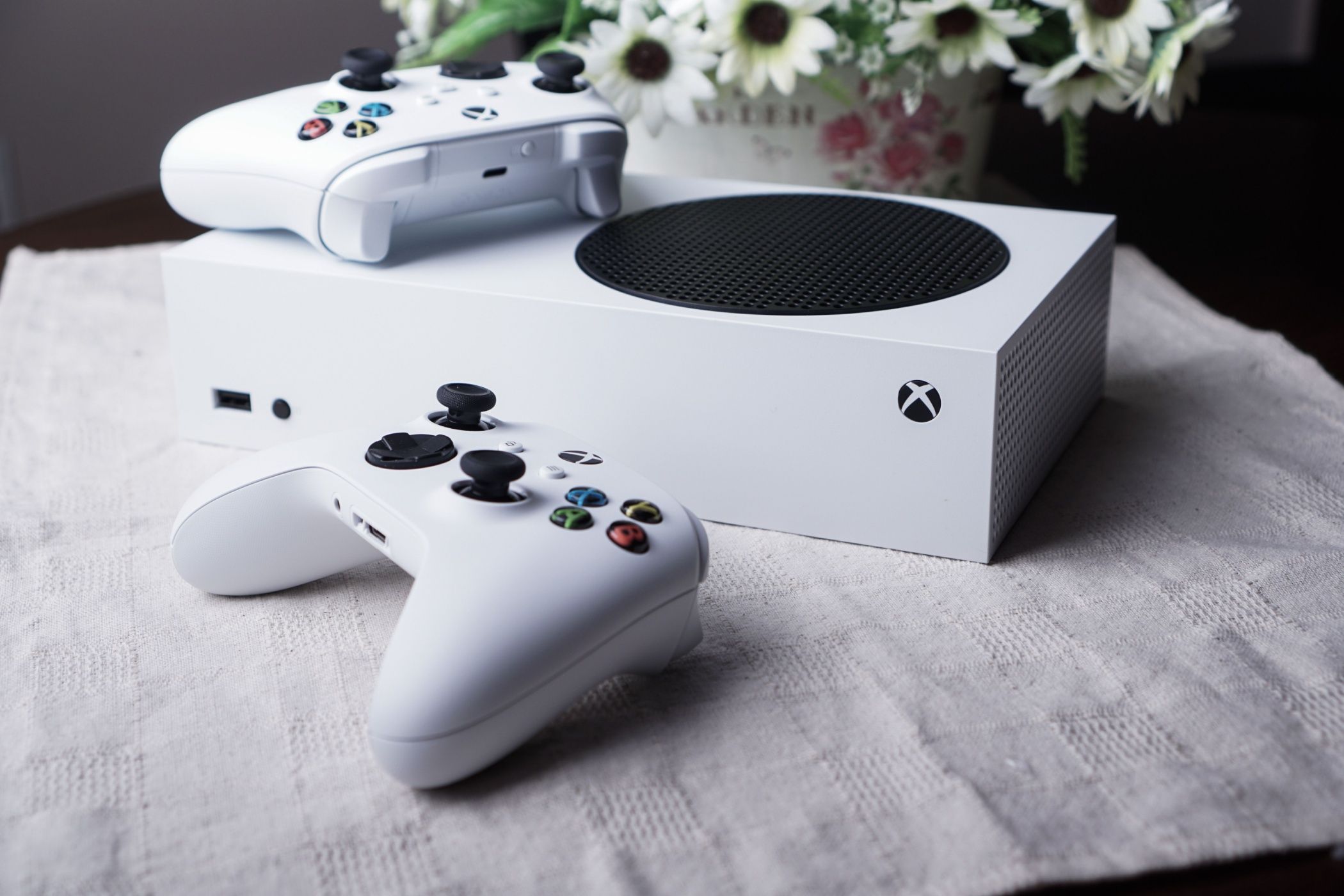 Um console Série S em uma mesa com dois controles e um vaso de flores.