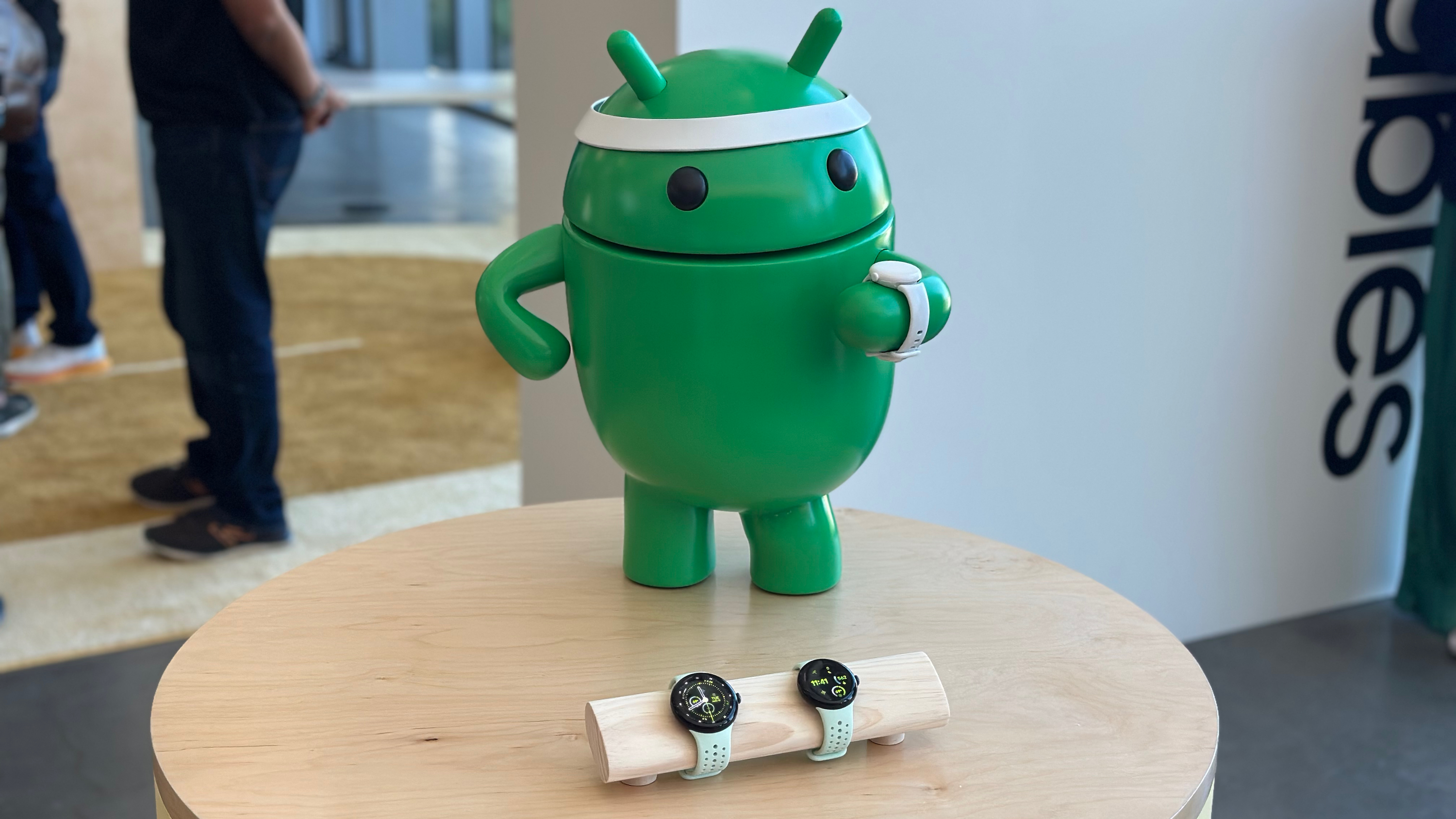 Uma estátua de Android usando uma faixa na cabeça e um boneco Pixel Watch 3, atrás de um display de Pixel Watch 3s.