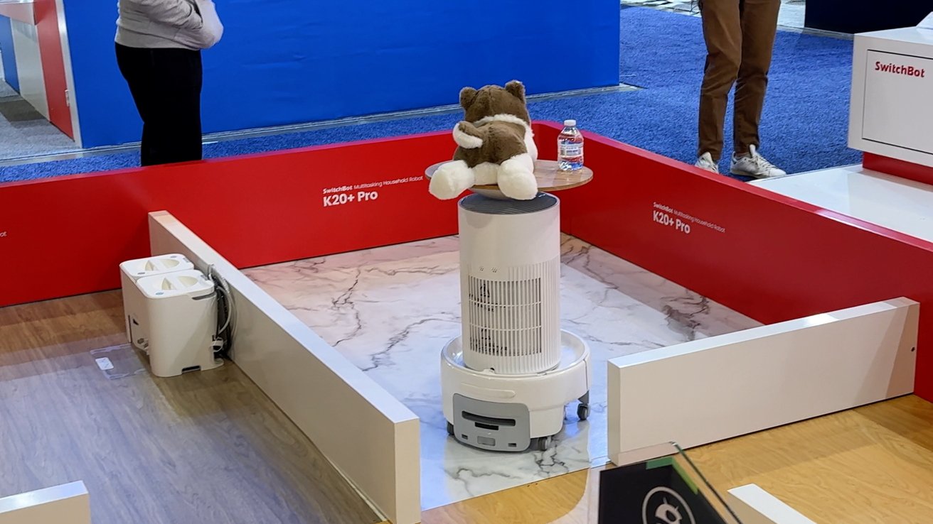 Robot displaying a stuffed animal and bottle atop, within a red enclosure labeled K20+ Pro. People are observing on blue carpet in background.