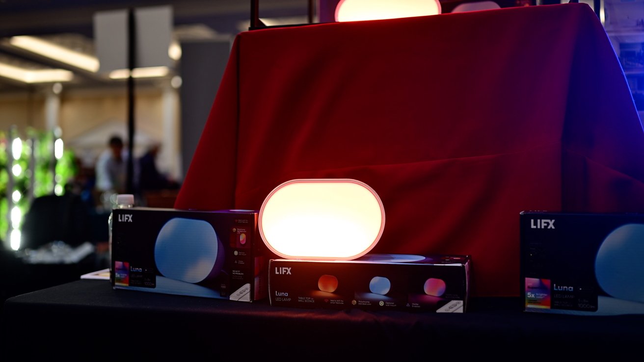 A bright oval lamp on a table with red cloth, surrounded by Lifx Luna LED lamp boxes, set in a dimly lit indoor environment.