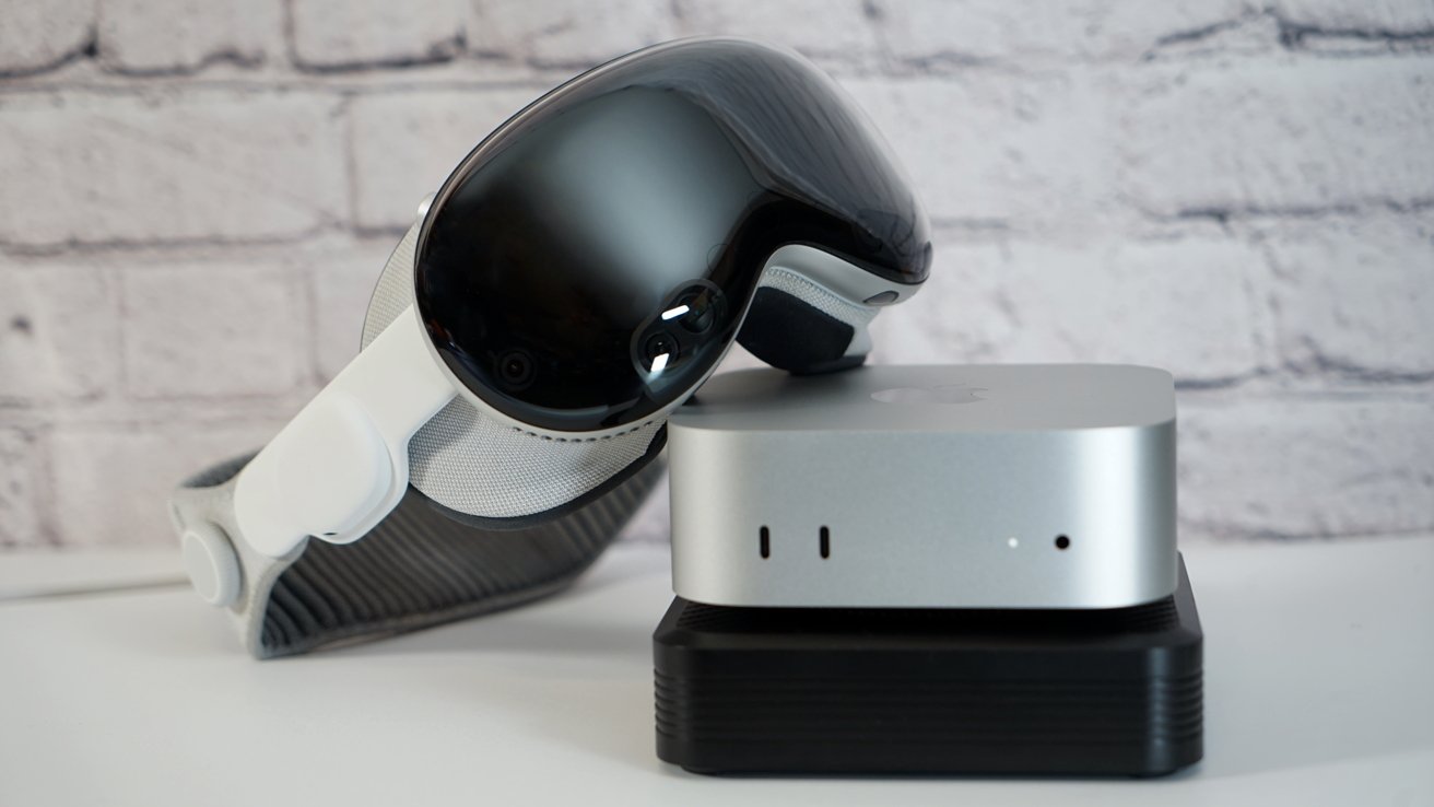 A sleek virtual reality headset rests against a compact silver computer on a dark base, set against a light brick wall background.