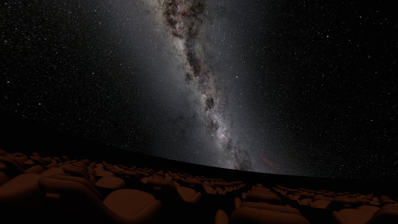 Interior do planetário com fileiras de assentos vazios voltados para uma projeção aérea da Via Láctea, cercada por um céu estrelado.