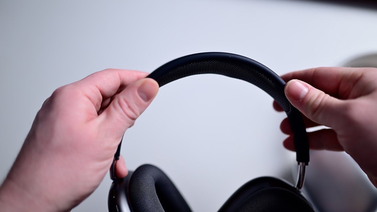 Mãos segurando fones de ouvido pretos, mostrando a faixa de cabeça contra um fundo simples.