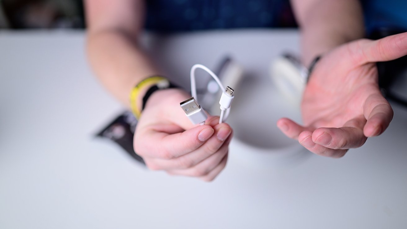 Uma mão segurando fones de ouvido com excesso de orelha branca e prata contra um fundo borrado, com um relógio visível no pulso.