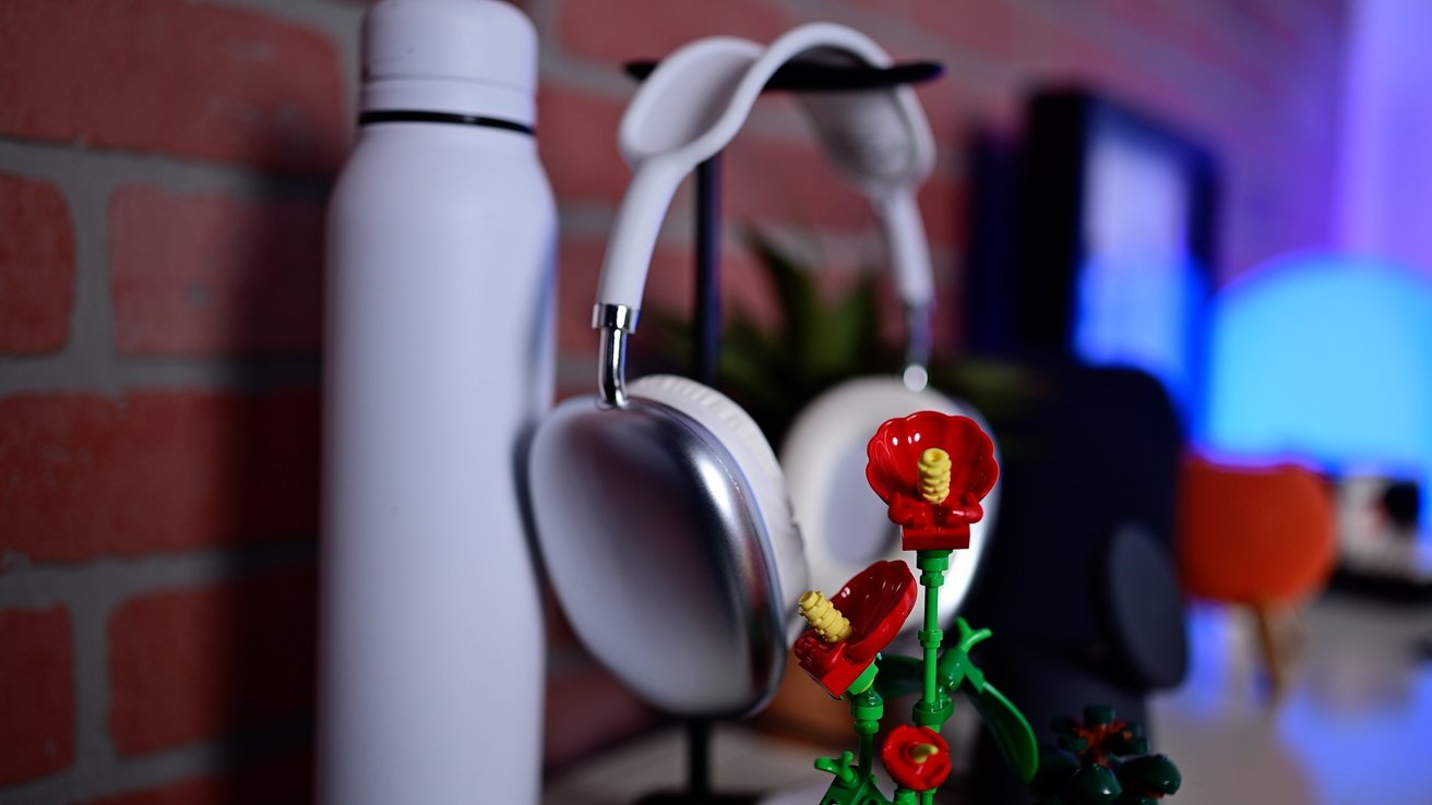 Fones de ouvido brancos, uma garrafa branca e um modelo de flores vermelhas em uma prateleira contra uma parede de tijolos.