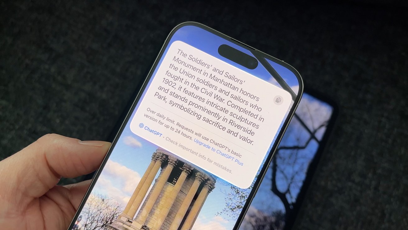 Mão segurando um smartphone exibindo texto sobre o monumento dos soldados e marinheiros, com uma imagem de fundo das colunas do monumento e galhos de árvores nuas.