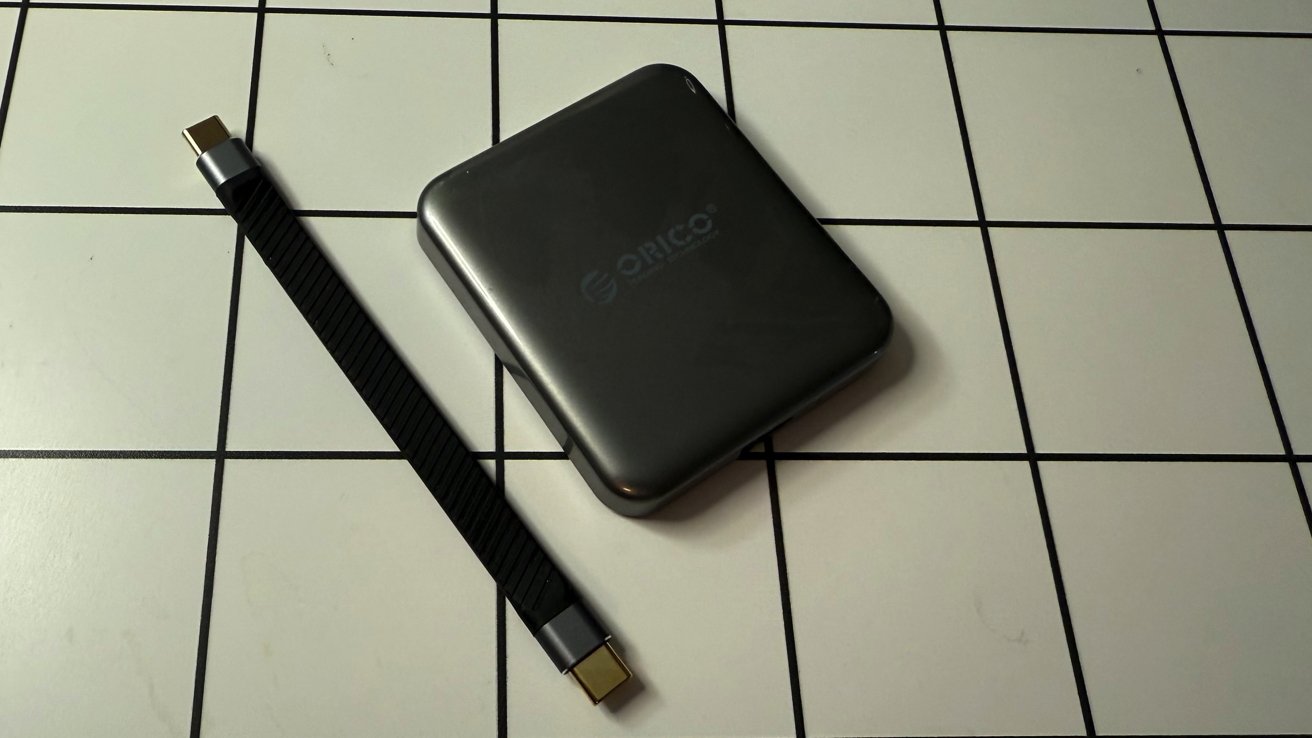 Um disco rígido externo quadrado preto com um logotipo fica ao lado de um cabo USB-C preto em uma superfície de azulejos.