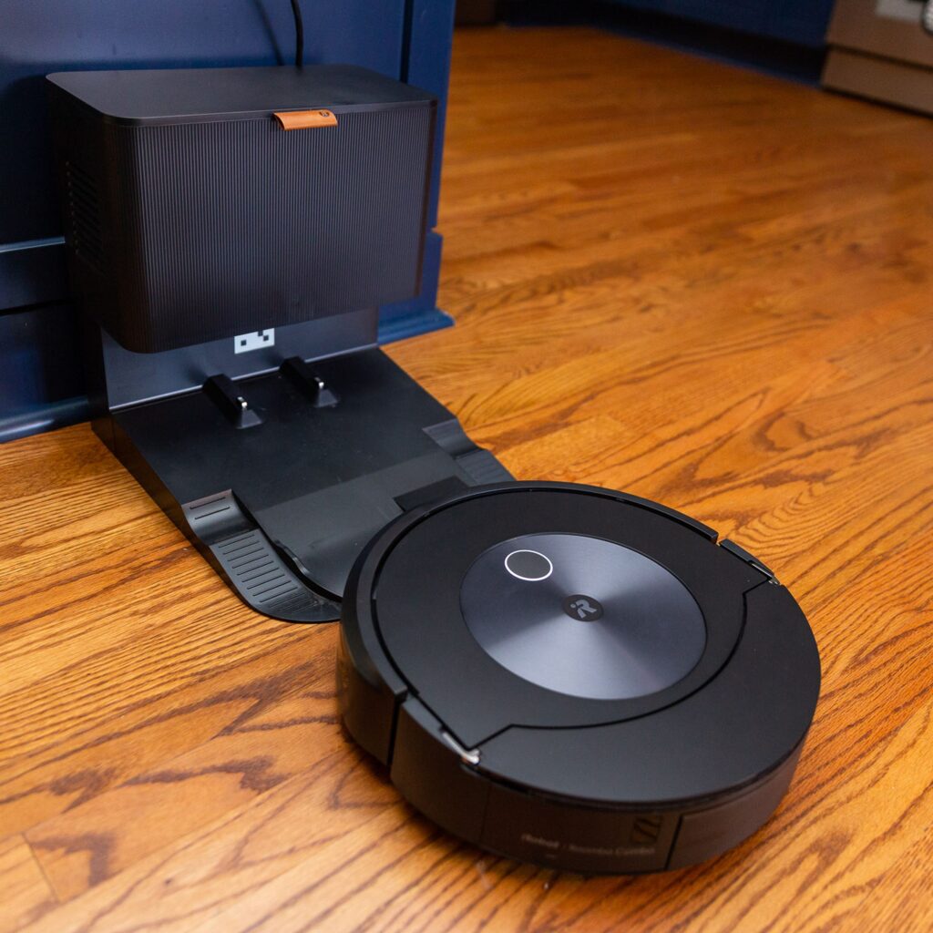 Foto da combinação do Roomba J7, aproximando-se de sua base automática. A base é do tamanho de uma pequena lata de lixo de cozinha.